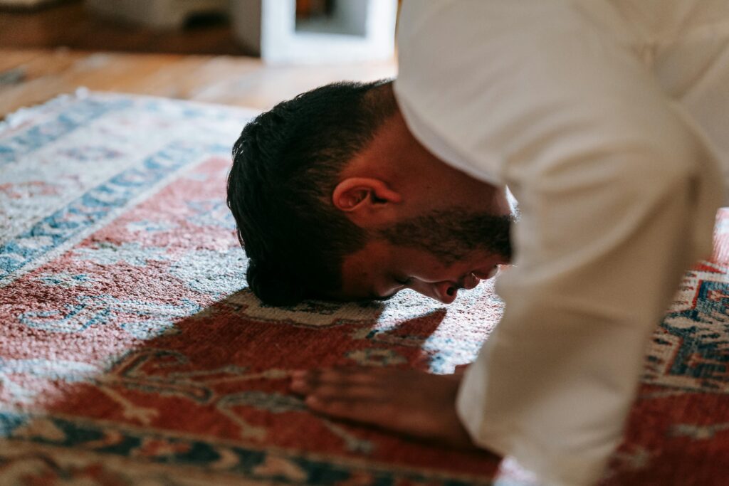 A Muslim man performing a prayer ritual inside on a traditional rug.