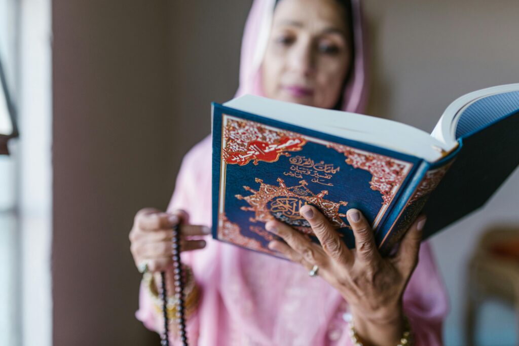 A woman in traditional attire reads the Quran indoors, holding prayer beads.